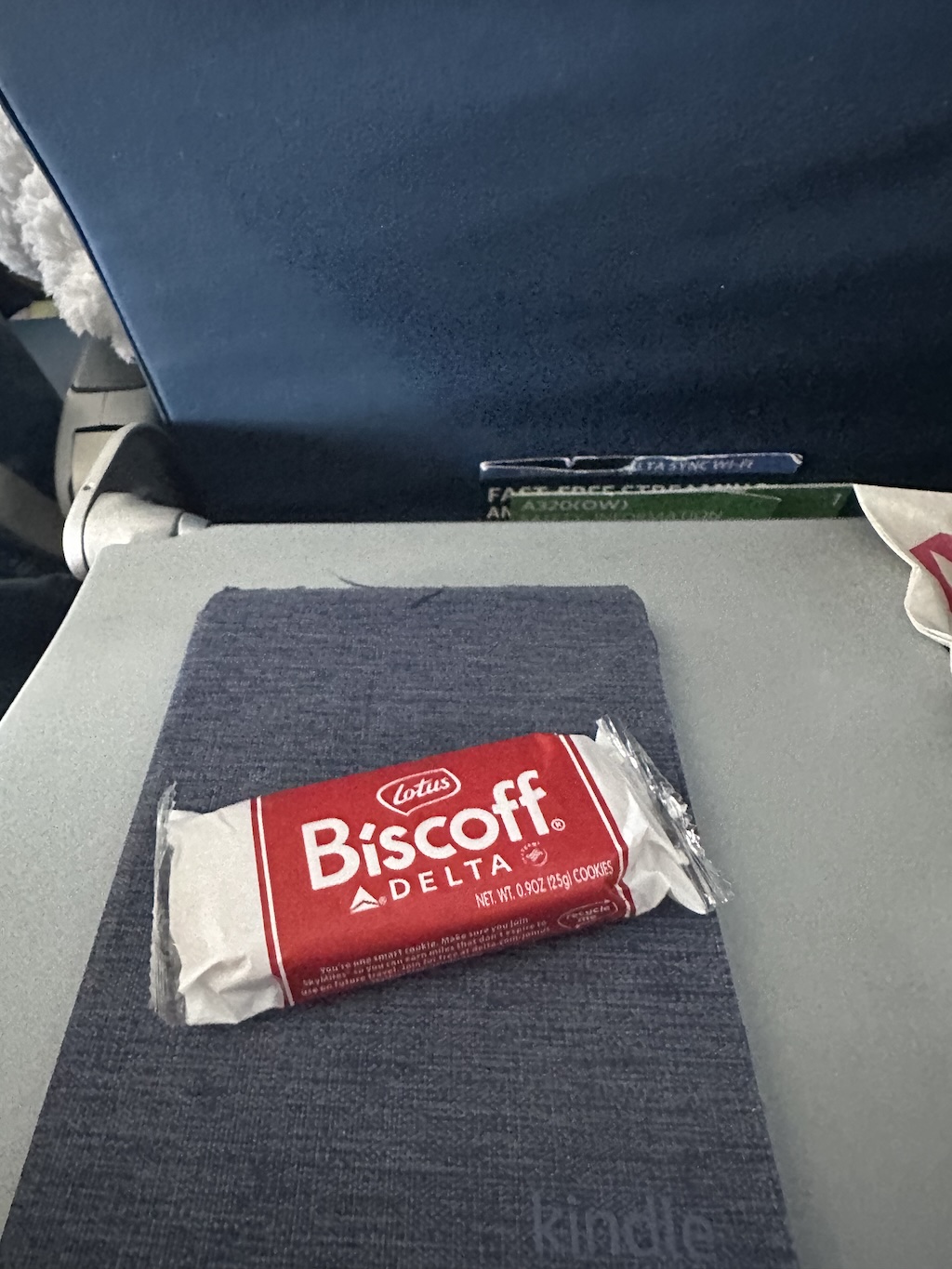 A package of Lotus Biscoff cookies with Delta branding is placed on a blue fabric cover, which appears to be a Kindle case, atop an airplan tray table. The airplane seatback and a seat pocket containing materials are visible in the background.