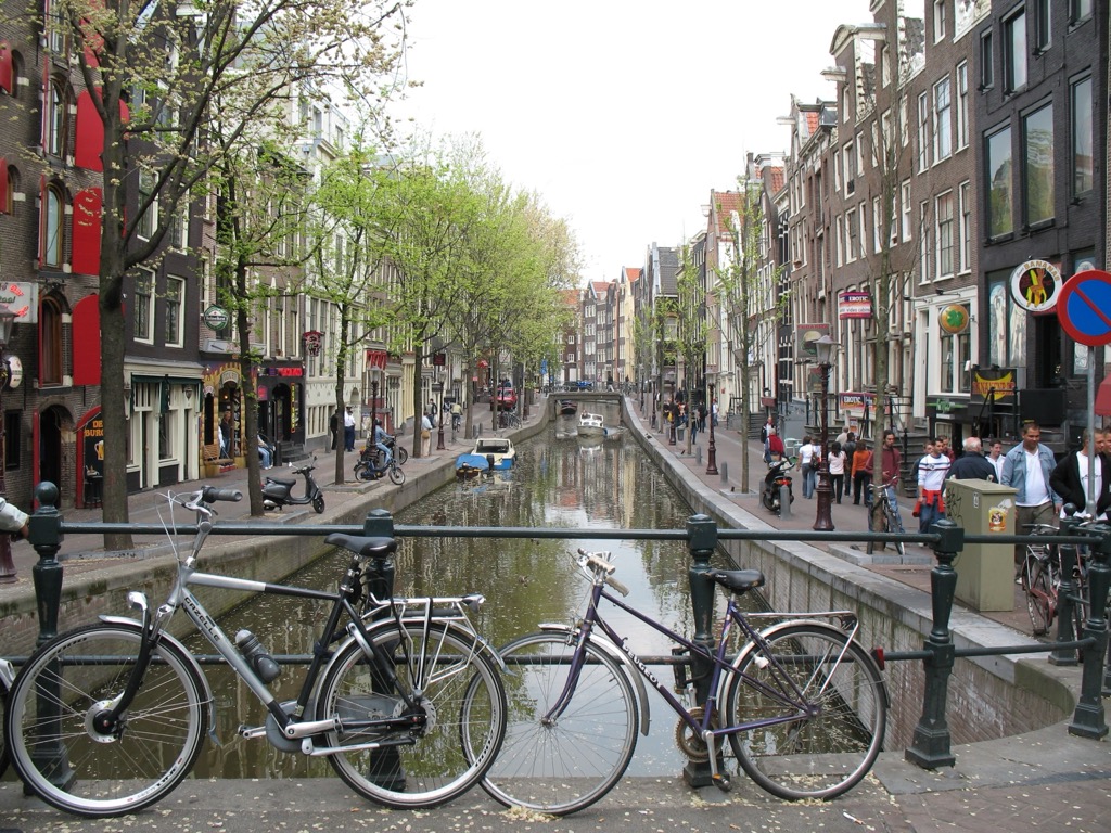 Two bicycles leaning against the black iron railing of an Amsterdam canal bridge, with a tree-lined canal, gabled houses, and pedestrians stretching into the distance.