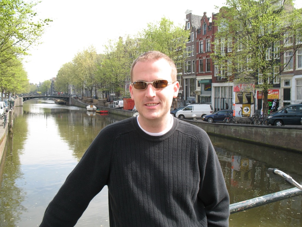 Brian, in a black sweater and sunglasses, stands on a bridge over an Amsterdam canal lined with trees, gabled houses, parked cars, and bicycles; a red 'Sexy Amsterdam' sign is visible on the right.