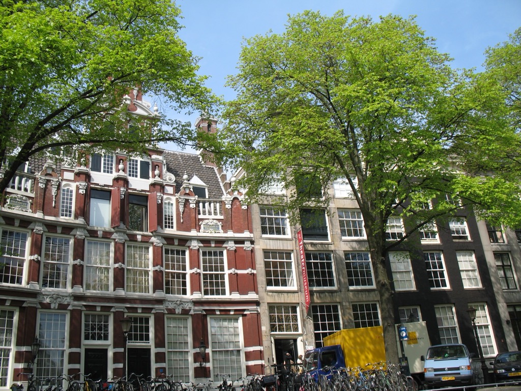 A row of ornate Amsterdam canal houses with red-and-white brick facades, white trim, and stepped gables, framed by bright green spring trees; bicycles and vans parked below.