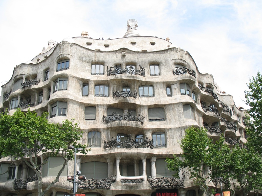 Gaudí's La Pedrera (Casa Milà) in Barcelona, an undulating pale stone façade with wavy balconies of twisted wrought iron and a sculpted rooftop, framed by green trees.