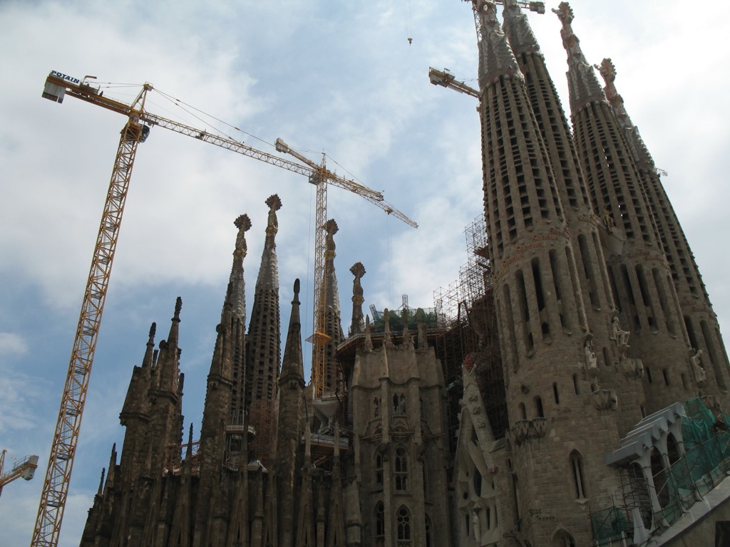 The Sagrada Família in Barcelona under construction, its stone spires and pinnacles surrounded by tall yellow tower cranes against a partly cloudy sky.