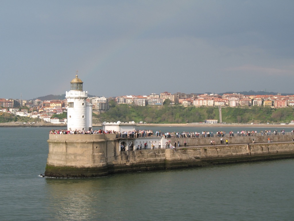Crowds of people line a stone breakwater topped by a white lighthouse at the mouth of Bilbao's harbor, with the town and green hills across the water under a hazy sky.