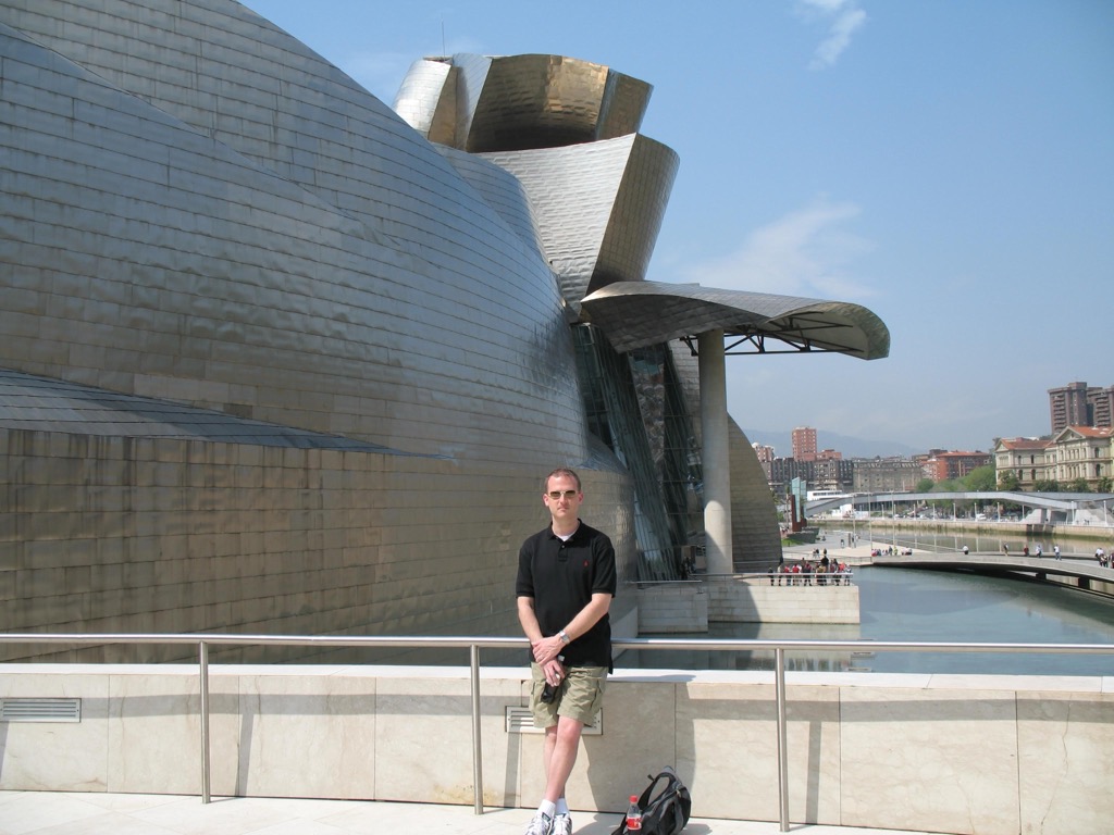 Brian, in a black polo and khaki shorts, stands at a stone railing in front of Frank Gehry's Guggenheim Museum Bilbao, its curving titanium panels gleaming above the river.
