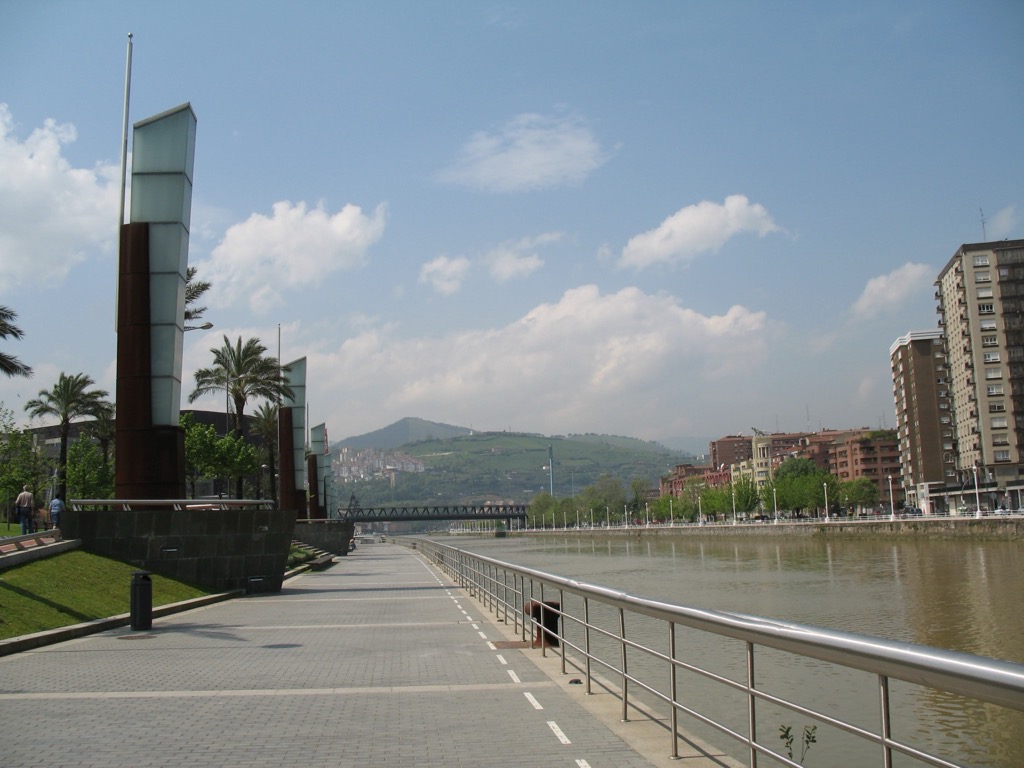 A wide tiled riverside promenade in Bilbao beside the Nervión river, with palm trees, tall modern sculptures, apartment blocks, and green hills rising under a blue sky.