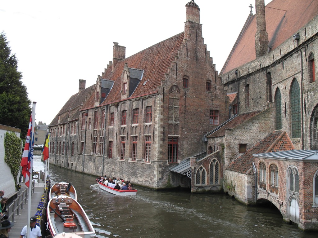 A Brugge canal lined with medieval brick buildings and stepped gables; a small open tour boat full of passengers glides along the green water under an overcast sky.