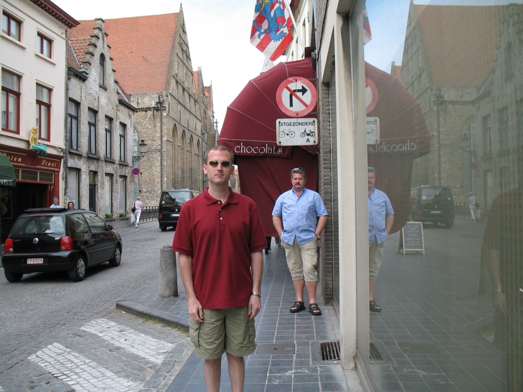 Brian, in a dark red polo and cargo shorts, stands on a cobbled Brugge street in front of a shop with a burgundy awning reading 'chocolats'; a Belgian flag and old stone buildings behind him.