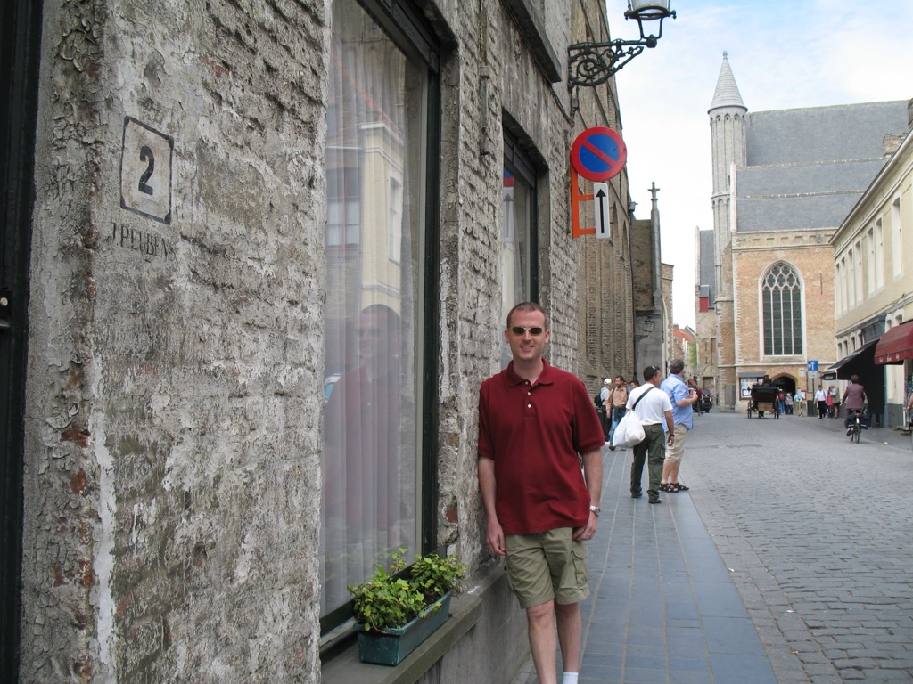 Brian, in a dark red polo, leans against a weathered whitewashed brick wall on a Brugge street; a Gothic church with a tall turret rises in the background beyond tourists on the cobbles.