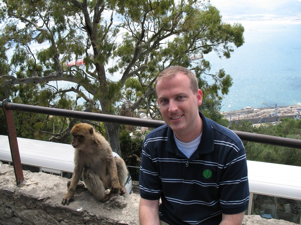 Brian, in a navy striped polo, crouches beside a Barbary macaque perched on a stone wall atop the Rock of Gibraltar, with the harbor and bay far below through the trees.