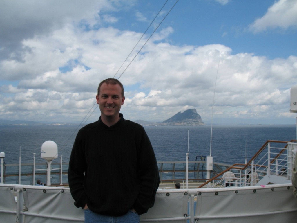 Brian, in a black sweater, stands smiling on the ship's top deck behind the white railing and a satellite dome; the Rock of Gibraltar rises across the water under a cloudy sky.