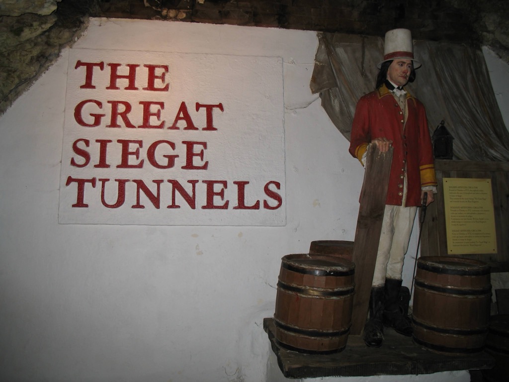Inside the Great Siege Tunnels of Gibraltar: a whitewashed rock wall painted 'THE GREAT SIEGE TUNNELS' in red, beside a uniformed red-coated soldier mannequin standing on wooden barrels.
