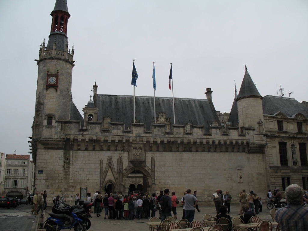 The fortified Gothic-Renaissance Hôtel de Ville of La Rochelle, with a tall clock-and-bell tower, crenellated stone walls, and French and EU flags; a crowd gathers at the entrance.