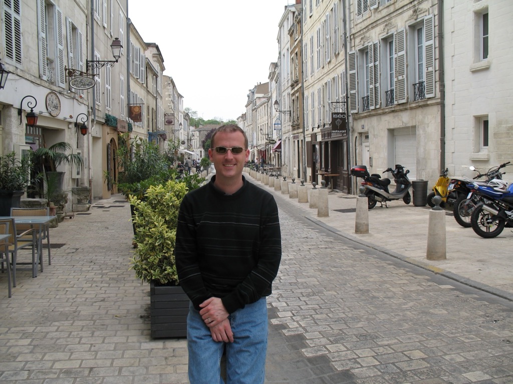 Brian, in a striped sweater and sunglasses, stands on a cobbled pedestrian street in La Rochelle lined with pale stone buildings, shop signs, café tables, and parked scooters.