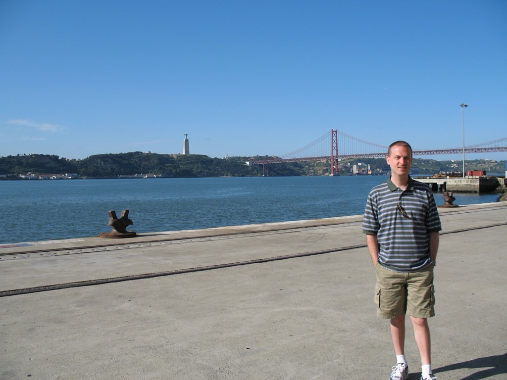 Brian, in a striped polo, stands on a broad concrete waterfront with the red 25 de Abril suspension bridge and the hilltop Cristo Rei statue across the Tagus river behind him.