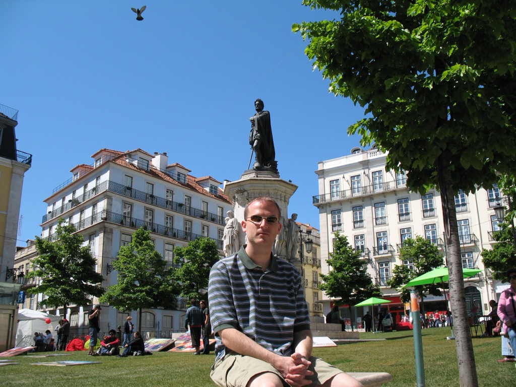 Brian, in a striped polo and sunglasses, sits on the grass in Praça Luís de Camões with the Camões monument and pale apartment buildings rising behind him under a bright blue sky.