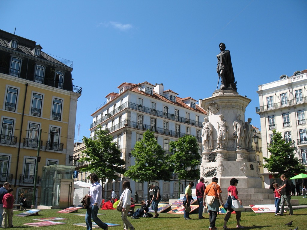 Lisbon's Praça Luís de Camões, a plaza with the bronze statue of Luís de Camões atop a stone pedestal ringed by smaller figures, surrounded by yellow and white buildings; people cross the square in the sun.