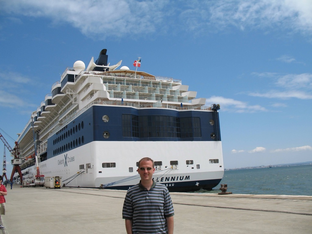 Brian, in a striped polo, stands on the quay in front of the docked Celebrity Millennium, whose blue-and-white hull and lifeboats tower behind him under a bright sky.