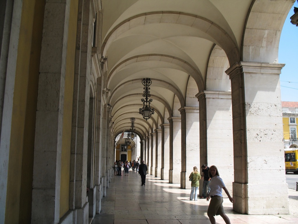 A long vaulted stone arcade at Lisbon's Praça do Comércio, with repeating arches, hanging lanterns, and yellow buildings; a woman walks through the light on the right and a yellow tram passes beyond.