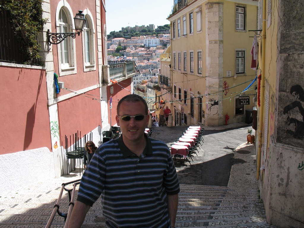 Brian, in a striped polo and sunglasses, stands on a steep stepped Lisbon street lined with pink and yellow buildings and café tables; the hilltop Castelo de São Jorge is visible across the city behind him.