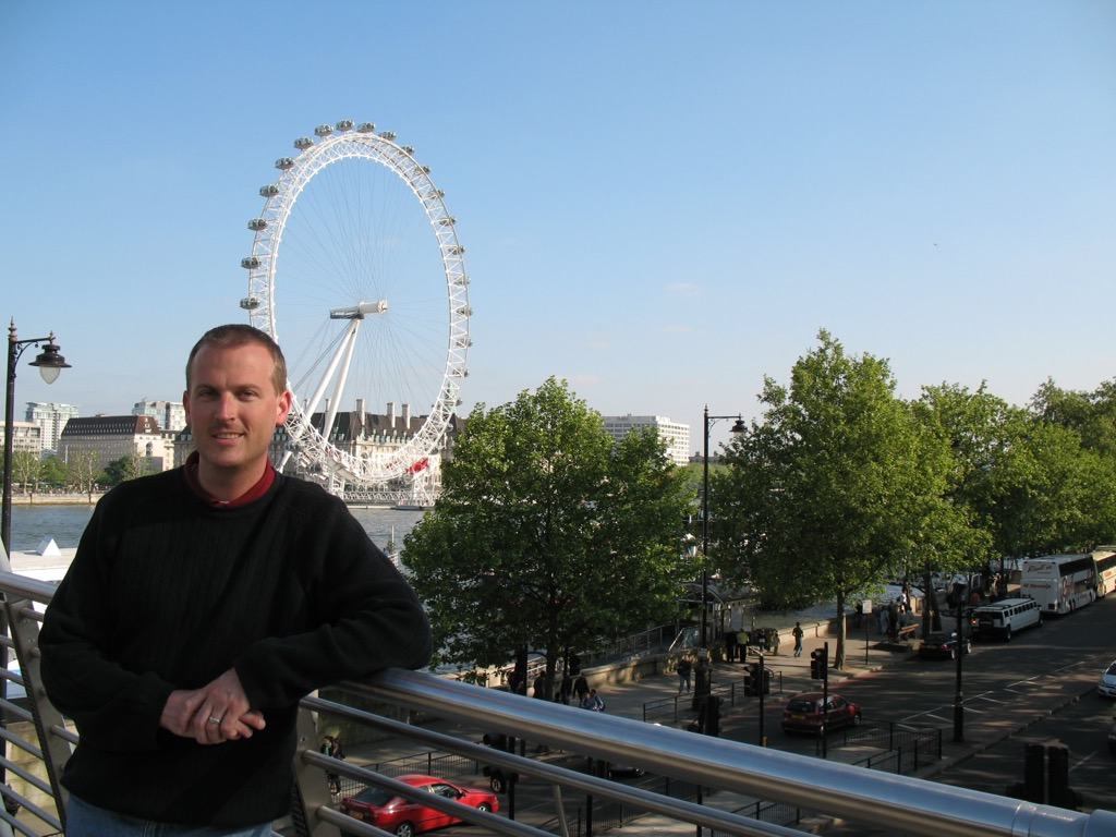 Brian, in a black sweater, leans on a riverside railing with the London Eye's white observation wheel rising behind him across the Thames, trees and a tour bus along the embankment.