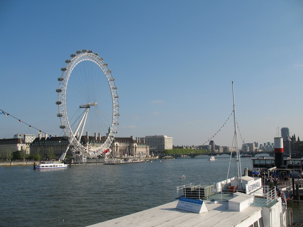 The London Eye seen across the River Thames under a clear blue sky, with sightseeing boats moored along the near bank and a bridge in the distance.