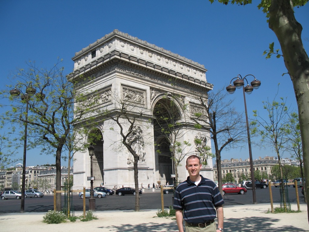 Brian, in a blue-striped polo, stands on a tree-lined plaza with the Arc de Triomphe and its carved reliefs behind him and cars circling the monument.