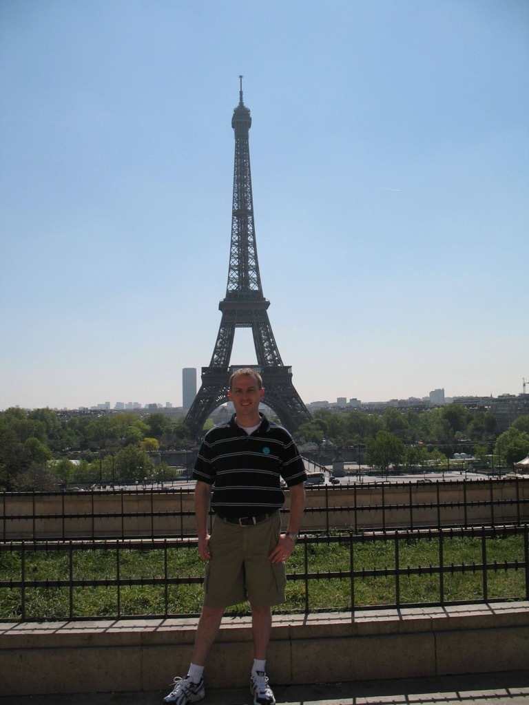 Brian, in a black striped polo and khaki shorts, stands smiling on the Trocadéro terrace with the Eiffel Tower rising behind him against a clear blue sky.