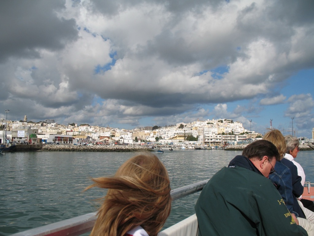 Tangier's white hillside town seen across the harbor from a tender boat under a dramatic cloudy sky; passengers sit at the rail in the foreground, one with windblown hair.