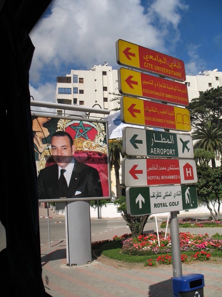 A cluster of Moroccan road signs in Arabic and French (Cité Universitaire, Aéroport, Hôpital Mohammed V, Royal Golf) beside a large billboard portrait of King Mohammed VI, with palm trees and white buildings behind.