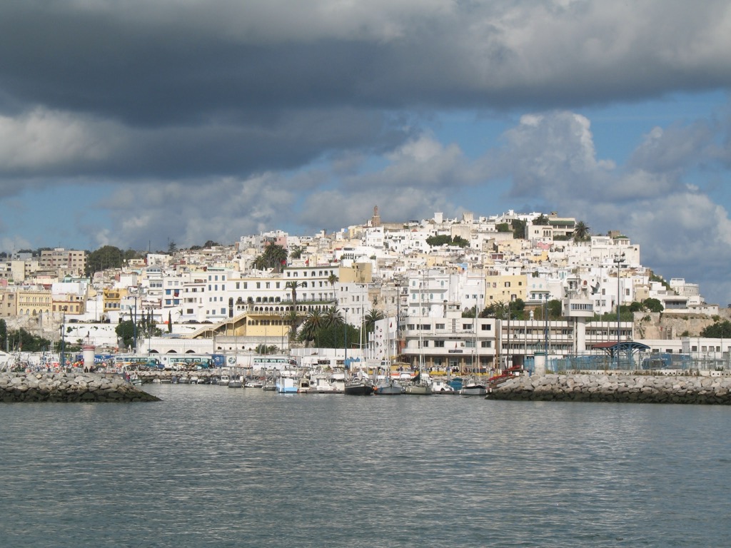 Tangier's whitewashed medina climbs a hillside above the harbor and its stone breakwater, boats moored below, under a sky of broken storm clouds.