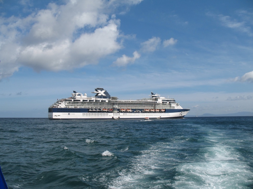 The Celebrity Millennium at anchor on open blue water under scattered clouds, seen from a small tender boat whose wake curves across the foreground.