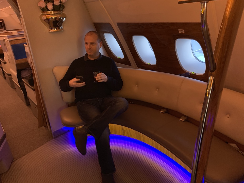 A man in a dark navy sweater and jeans sits cross-legged on the curved beige leather banquette of the onboard lounge on an Emirates A380, holding a phone in one hand and a short glass in the other, with wood-paneled walls, three oval windows, a brass vase of pink roses mounted on the wall, a polished brass pole in the foreground, and blue LED lighting glowing along the base of the seat