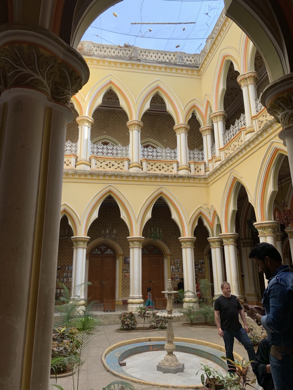 Inner courtyard of Bangalore Palace, framed by a carved stone column in the foreground: two stories of pale yellow pointed Gothic arches trimmed in orange and cream, white columns with gilded capitals, ornate white latticework railings on the upper gallery, framed photographs on the walls below, and a small tiled fountain surrounded by potted plants. A man in a black T-shirt stands in the courtyard while another man looks at his phone at right, under a partly open sky strung with wires