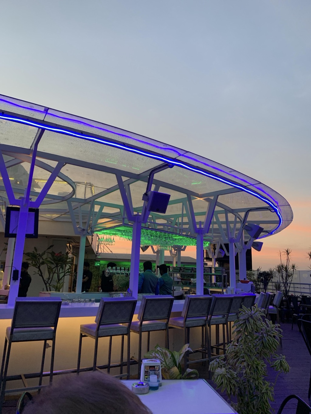 Rooftop bar in Bengaluru at dusk, with a curved translucent canopy on white steel supports edged in glowing blue LED strip lighting, green-lit racks of hanging wine glasses above the bar, bartenders at work, a row of empty gray bar stools along the lit counter, potted palms, and an orange and pink sunset on the horizon