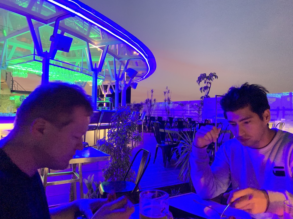 Two men eating dinner at a table on the same Bengaluru rooftop after dark, both lit in purple and blue from the LED-edged canopy behind them: a man in a dark T-shirt in the foreground at left and a man in a white G-Star sweatshirt at right, with a pint of beer on the table and empty tables, chairs, and potted plants across the deck under a hazy violet sky