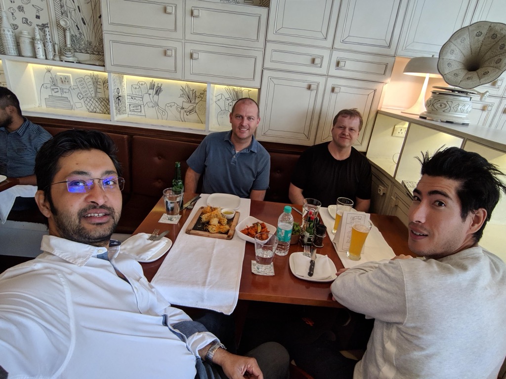 Selfie of four men around a restaurant table in Indiranagar, Bengaluru: a man in glasses and a white shirt in the foreground, a man in a blue polo and a man in a black T-shirt seated against a brown leather banquette, and a man in a gray sweatshirt at right. The table holds a wooden board of fried appetizers, a bowl of chicken, bottled water, and glasses of beer, and the cream-colored cabinets behind them are painted with whimsical line drawings, beside a lamp shaped like a gramophone horn