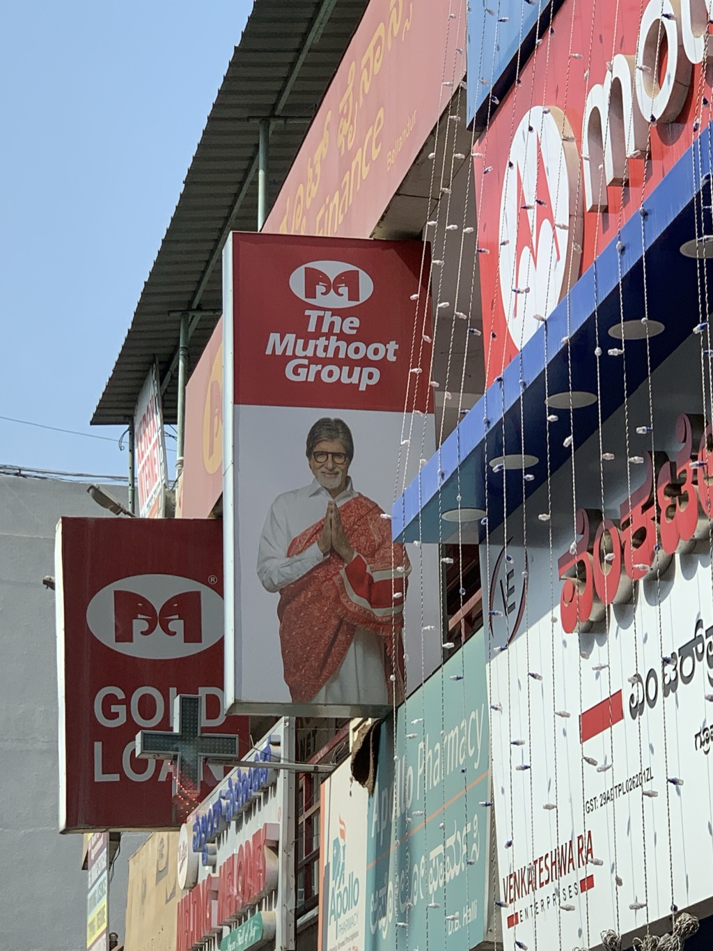 Crowded shop signage on a street in Bellandur, Bengaluru, under a clear blue sky: a red and white sign for The Muthoot Group featuring actor Amitabh Bachchan in a red shawl with his palms pressed together in greeting, a red Gold Loan sign with the Muthoot elephant logo, a green pharmacy cross, an Apollo Pharmacy sign, and a large sign for Venkateshwara Enterprises in Kannada and English script, with strings of small lights hanging across the storefronts