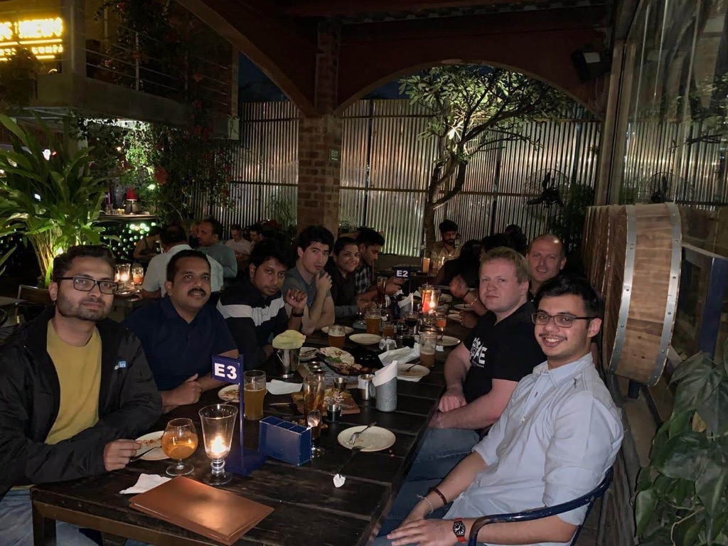 A group of about a dozen colleagues seated along a long wooden table at a dinner at an open-air brewpub in Bengaluru at night, many smiling at the camera, with glasses of beer and candles in glass holders on the table, a blue E3 table marker, a large wooden barrel at right, and corrugated metal walls, a brick arch, and potted plants behind them