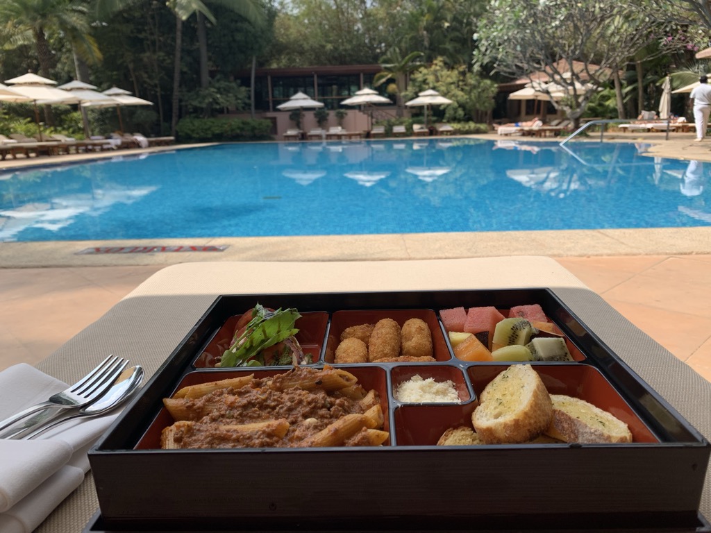 A black lacquered bento box lunch on a padded poolside lounger at The Leela Palace in Bengaluru, its red compartments holding penne in meat sauce, fried croquettes, a small salad, grated cheese, garlic bread, and watermelon, papaya, and kiwi, with silverware on a white napkin beside it and the bright blue pool, white umbrellas, and loungers under tropical trees in the background