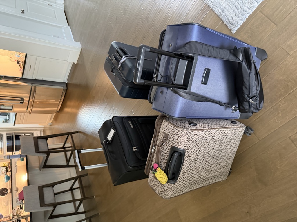 Four packed roller suitcases staged together on the hardwood floor of a kitchen at home — a black softside bag with a luggage tag, a dark hardside spinner, a tan patterned bag with a yellow pineapple tag, and a large navy softside bag — with bar stools, a stainless refrigerator, and white cabinetry in the background