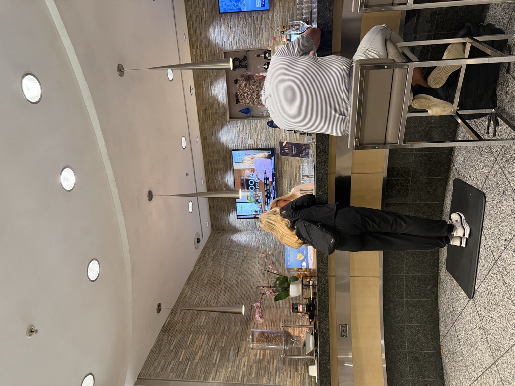 Interior of an airport lounge at RDU showing a curved granite bar with a stone-tiled wall behind it, three tapered pendant lights, recessed ceiling lighting, and wall-mounted televisions. A woman in black stands at the bar and a man in a striped shirt sits on a metal bar stool with a bag at his feet