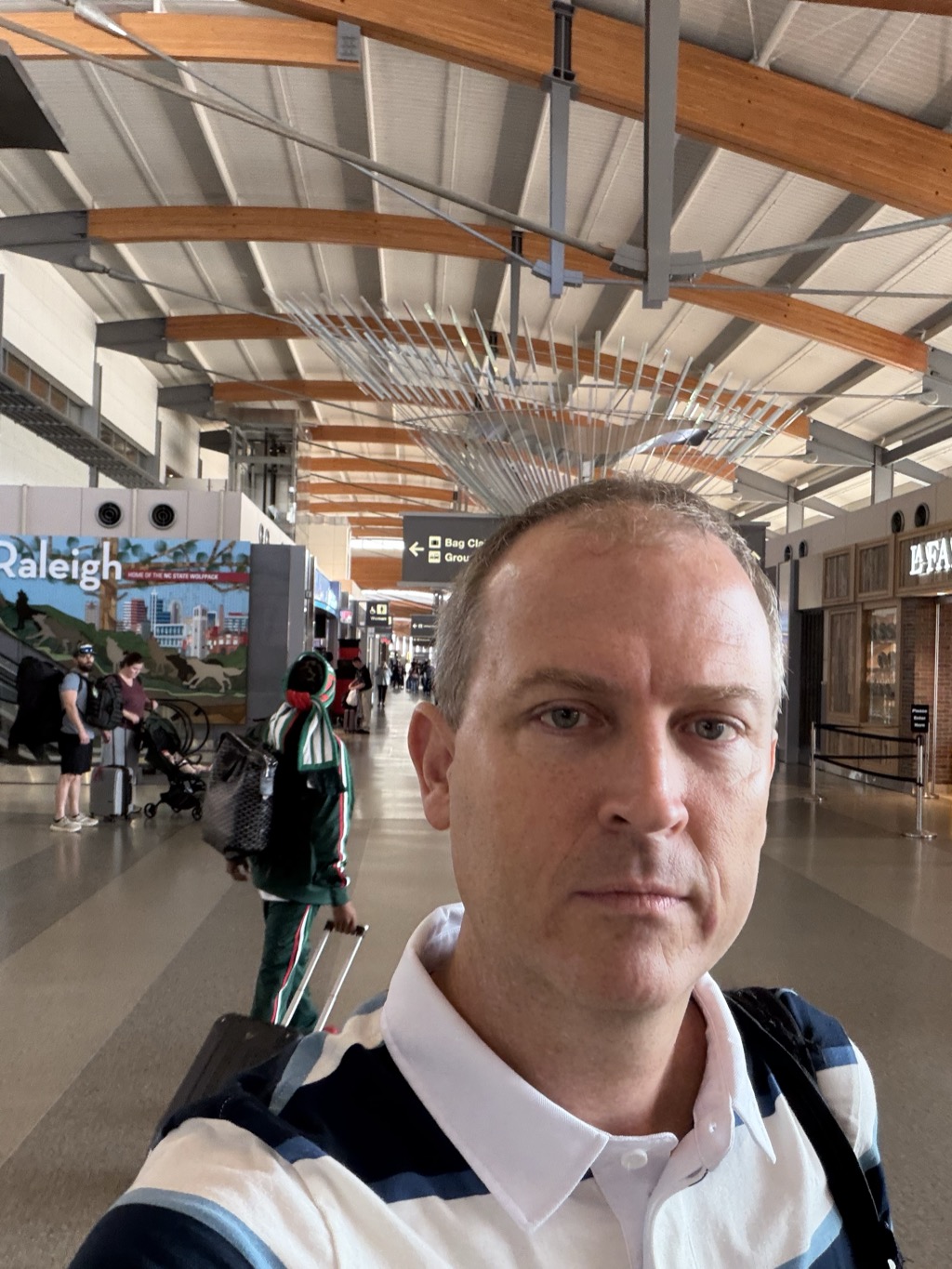 Selfie in the Raleigh-Durham International Airport terminal, wearing a navy and white striped polo with a backpack strap over one shoulder, standing beneath the terminal's curved wooden roof beams and spiked metal sculpture, with a colorful 'Raleigh — Home of the NC State Wolfpack' mural and other travelers with luggage in the background