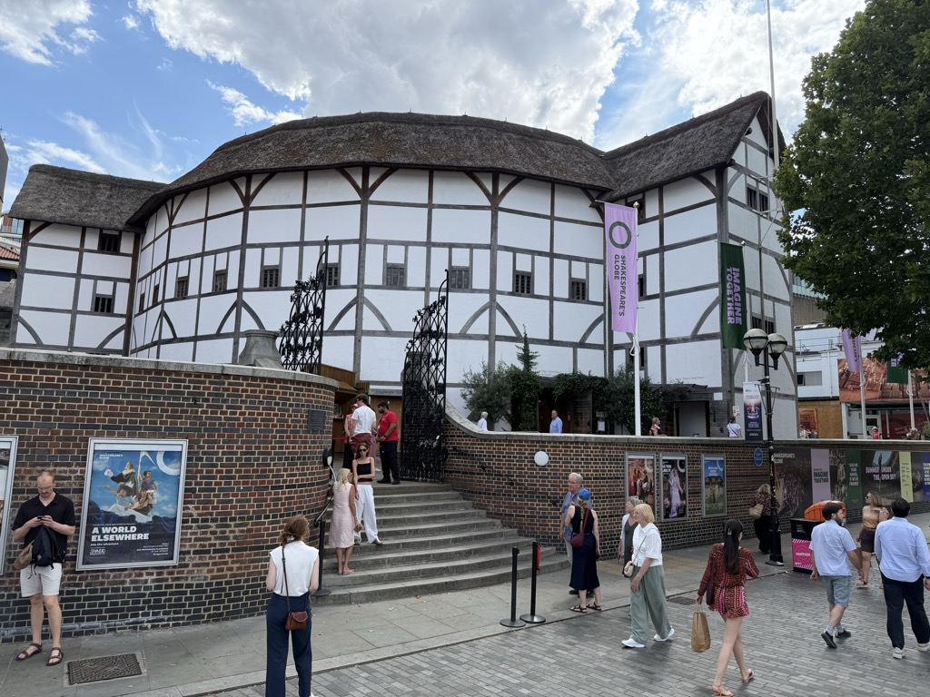 Shakespeare's Globe theatre in Bankside, London, a white timber-framed circular building with a thatched roof, photographed from the paved forecourt. Purple and green banners reading 'Shakespeare's Globe' and 'Imagine Together' hang from the facade, and a curved brick wall with a poster for the production 'A World Elsewhere' fronts the steps where visitors are entering. Pedestrians walk across the foreground on a partly cloudy afternoon
