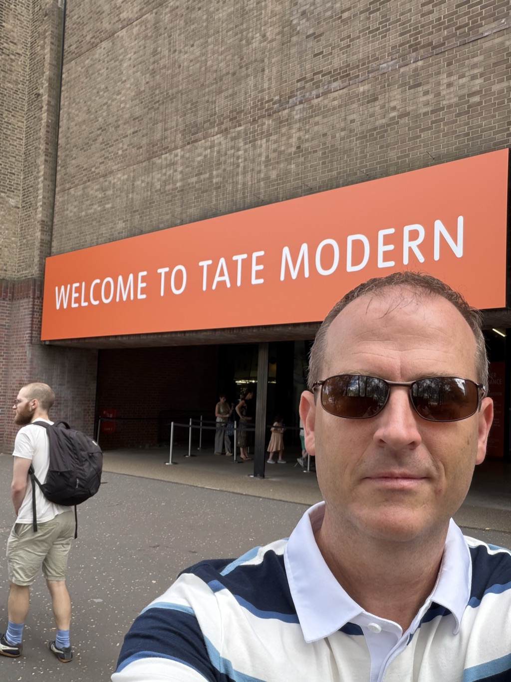 Selfie outside the Tate Modern in London, wearing sunglasses and a navy and white striped polo, standing beneath a large orange banner with white lettering reading 'Welcome to Tate Modern' mounted on the gallery's tan brick facade. A man with a black backpack walks past on the left and other visitors queue behind stanchions at the entrance