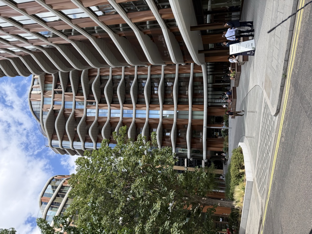 The Triptych Bankside residential towers seen from street level, with curving white balcony bands wrapping around terracotta and glass facades against a partly cloudy blue sky. At ground level people sit on wooden benches in a landscaped plaza with young trees and planters, and three men in business attire stand talking beside a Triptych information sign near the entrance