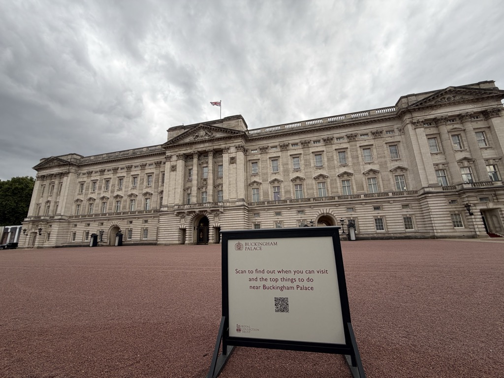 The east front of Buckingham Palace photographed from across the empty forecourt on an overcast day, showing the full symmetrical stone facade with its central pediment and the Union Flag flying from the roof. A Royal Collection Trust A-board in the foreground reads 'Buckingham Palace — Scan to find out when you can visit and the top things to do near Buckingham Palace' with a QR code