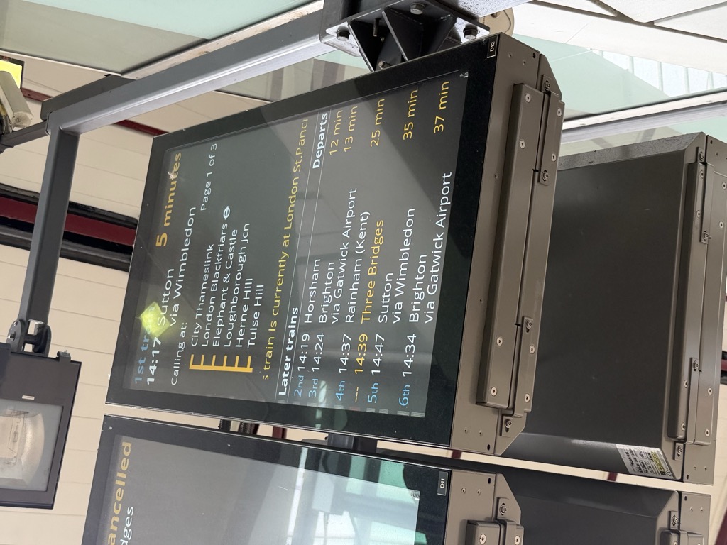 Overhead electronic departure board at Farringdon station showing the first train at 14:17 to Sutton via Wimbledon in 5 minutes, calling at City Thameslink, London Blackfriars, Elephant and Castle, Loughborough Junction, Herne Hill, and Tulse Hill, with a note that the train is currently at London St Pancras. Later trains listed include Horsham at 14:19, Brighton via Gatwick Airport at 14:24, and Rainham in Kent at 14:37. A cancelled notice is partly visible on the adjacent screen