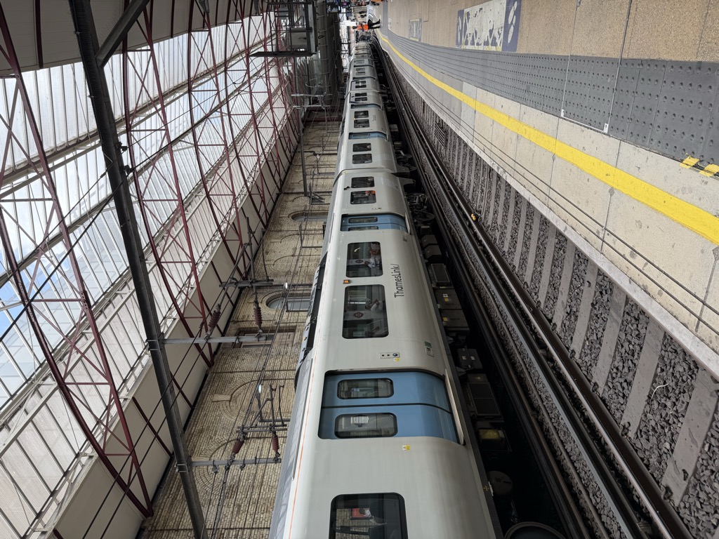 A white and blue Thameslink train stopped alongside a platform at Farringdon station, seen from the yellow platform edge line. The station has a curved glass and red steel roof over yellow brick walls with arched windows, and passengers are visible through the train windows with a worker in a hi-vis vest further down the platform