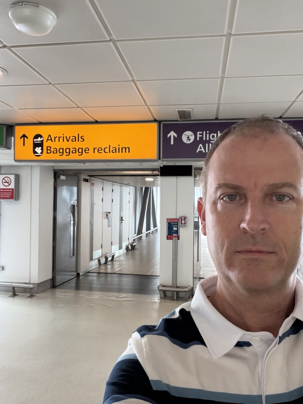 Selfie in a London Heathrow Airport corridor after landing, wearing a navy and white striped polo, standing beneath a yellow overhead sign reading 'Arrivals Baggage reclaim' with an up arrow and a purple sign for flight connections. A long empty white-walled walkway with a no-smoking notice and a red fire alarm point stretches into the background