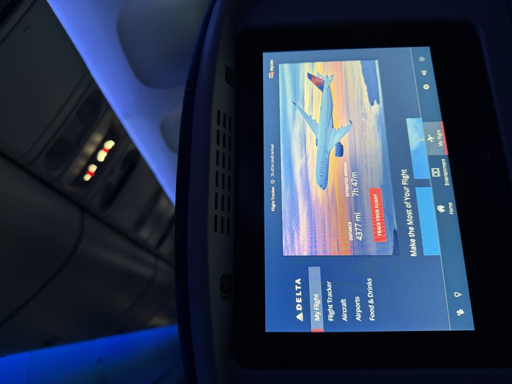 Delta seatback entertainment screen photographed from a darkened aircraft cabin, showing the My Flight page with an illustration of a Delta jet against a sunset sky, a distance of 4377 miles, an estimated arrival of 7 hours 47 minutes, and a red 'Track Your Flight' button. Overhead cabin lighting and the curved blue seatback of the row ahead are visible above the screen